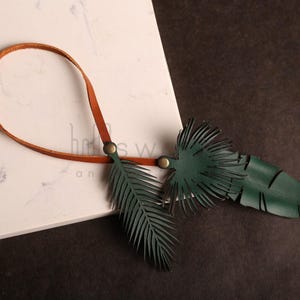 Pode incluir: Um cord&atilde;o de couro castanho com dois pingentes de couro verde em forma de folhas de palmeira. Os pingentes s&atilde;o presos ao cord&atilde;o com argolas de metal e t&ecirc;m uma superf&iacute;cie texturizada.