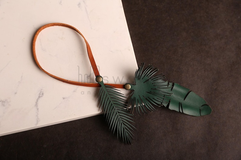 Pode incluir: Um cord&atilde;o de couro castanho com dois pingentes de couro verde em forma de folhas de palmeira. Os pingentes s&atilde;o presos ao cord&atilde;o com argolas de metal e t&ecirc;m uma superf&iacute;cie texturizada.
