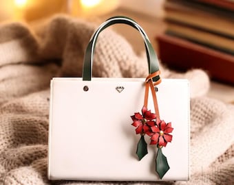 Puede incluir: Bolso de mano de cuero blanco con asa verde y blanca y correa de cuero marrón con dos abalorios de flores rojas de cuero. El bolso está sobre una manta de punto blanca.