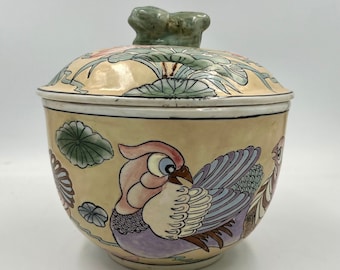 May include: A ceramic jar with a lid featuring a bird and floral design. The jar is cream colored with green, pink, purple, and brown accents. The lid has a frog finial.