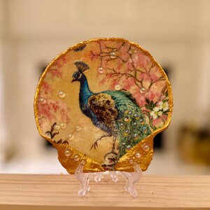 May include: A decorative seashell dish featuring a peacock illustration. The peacock is depicted in shades of blue and green, set against a backdrop of pink blossoms. The shell's edge is trimmed with gold paint and adorned with small, clear embellishments.