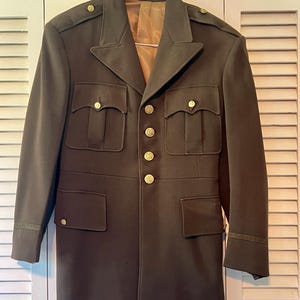 May include: A vintage military uniform jacket in a dark green color with gold buttons and a brown lining. The jacket has a pointed collar, two flap pockets on the chest, and two patch pockets on the lower front. The sleeves have a gold trim.