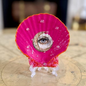 May include: A vibrant pink seashell art piece featuring a detailed eye within a crescent moon design. The shell is adorned with clear, sparkling embellishments and rests on a clear stand, creating a unique decorative item.