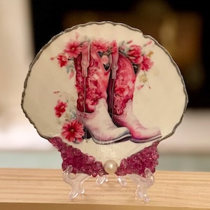 May include: A decorative, shell-shaped piece featuring a pair of pink cowboy boots with floral accents. The boots are set against a cream background and surrounded by pink flowers. The piece is supported by a clear stand.