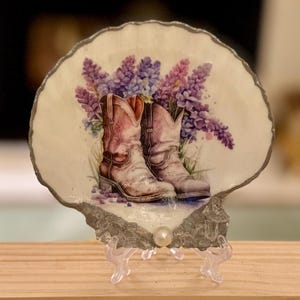 May include: A decorative seashell with a painted image of pink cowboy boots and purple flowers. The shell is mounted on a clear plastic stand with a pearl accent. The shell has a silver-colored edge.
