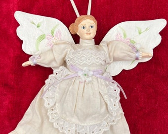 May include: A white porcelain angel ornament with painted wings, a white dress with lace trim, and a purple ribbon. The angel has brown hair and a painted face.