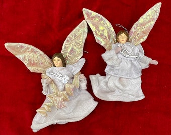 May include: Two white angel ornaments with iridescent wings. One angel holds a silver trumpet and the other holds a silver guitar. Both angels are wearing white dresses with gold trim.