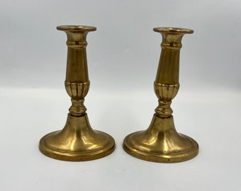 May include: Two gold metal candle holders with a fluted design. The candle holders are on a white surface.
