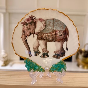 May include: A decorative seashell plate featuring an elephant design with floral accents and a gold trim. The elephant is adorned with a floral headdress and decorative coverings. The plate is displayed on a clear stand with green and gold accents.