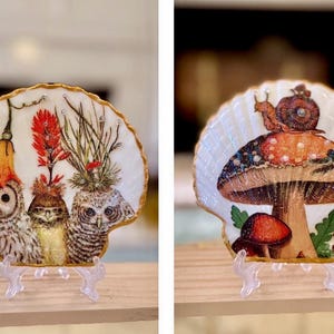 May include: Two decorative seashell art pieces. One features three owls with floral accents, the other depicts a mushroom with a snail and dragonfly. Both have gold trim and are displayed on clear stands.