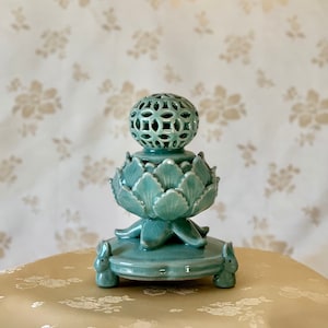 May include: A teal blue ceramic incense burner shaped like a lotus flower with a decorative, openwork sphere on top. The burner has three rabbit-shaped legs.