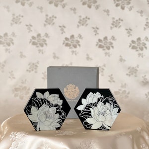 May include: Two black hexagonal coasters with white floral designs. The coasters feature a white lotus flower design on a black background. The coasters are made of a smooth, polished material.