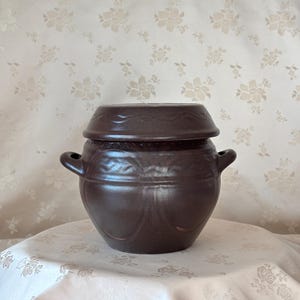May include: A dark brown ceramic jar with a matching lid and two side handles. The jar features a rounded body with embossed decorative patterns around the upper section and the lid, showcasing a smooth, matte finish throughout the exterior.