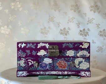 May include: A purple wooden jewelry box with two drawers, decorated with mother of pearl inlay and floral designs. The box has a silver lock and a green tassel.