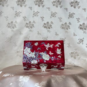 May include: A red jewelry box with a butterfly-shaped clasp. The box is adorned with mother-of-pearl floral and butterfly designs. The background features a floral pattern.