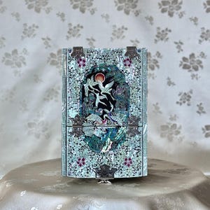 May include: A white and blue jewellery box with a mother-of-pearl inlay design. The box features a floral pattern and a scene of birds in flight. The box has two compartments and a hinged lid.