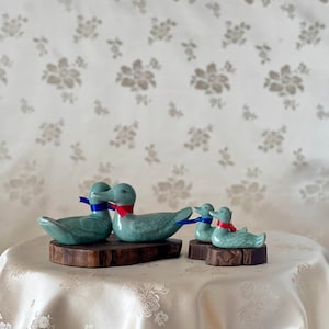 Pode incluir: Quatro figuras de patos de cerâmica em vários tamanhos, cada uma com uma fita azul ou vermelha amarrada ao pescoço. Os patos são verde claro e repousam sobre pequenas bases de madeira escura. O fundo apresenta um padrão floral.