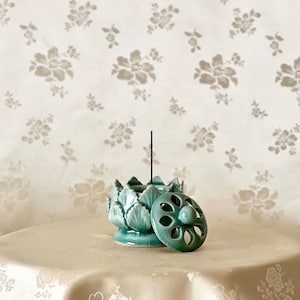 Op de afbeelding: Een tealblauwe keramische wierookbrander in de vorm van een lotusbloem met een deksel. De wierookbrander staat op een wit tafelkleed met een bloemenpatroon.