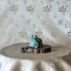Puede incluir: Una figurita de rana de cerámica azul claro sentada sobre una base de madera. La rana tiene una boca ancha y ojos grandes.