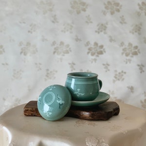 Könnte beinhalten: Ein Teekannen-Set aus blaugrüner Keramik mit Deckel. Tasse und Untertasse haben ein zartes Blumenmuster. Der Deckel hat ein kreisförmiges Design mit einer erhabenen Mitte.