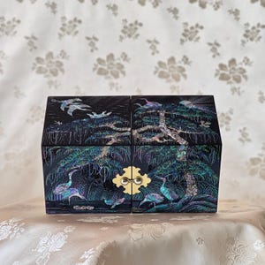 May include: A black jewellery box with a gold clasp, featuring an intricate mother-of-pearl inlay design. The design depicts cranes, trees, and foliage. The box is rectangular with a hinged lid, showcasing traditional artistry.