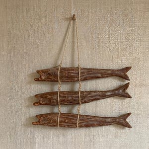 Puede incluir: Tres esculturas de peces marrones colgadas de una cuerda de fibra natural. Los peces están dispuestos verticalmente, cada uno atado a la cuerda con hilo. Los peces tienen la boca abierta y aletas detalladas.