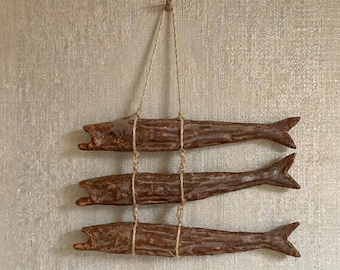 May include: Three brown fish sculptures hanging from a natural fiber string. The fish are arranged vertically, each tied to the string with twine. The fish have open mouths and detailed fins, creating a rustic, nautical aesthetic.