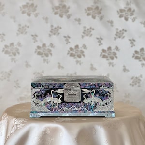 May include: A black wooden jewellery box with a hinged lid and a silver latch. The box is decorated with iridescent mother-of-pearl inlay depicting cranes and flowers. The box is sitting on a white fabric surface.