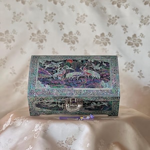 May include: A rectangular, iridescent jewelry box with a detailed inlaid design featuring cranes, birds, and a sun. The box has a silver clasp and a purple pen with decorative beads. The box is on a cream-colored surface.
