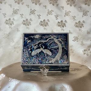 May include: A dark blue jewellery box with a mother-of-pearl inlay showing cranes, trees, and a full moon. The box has a silver butterfly clasp and is displayed on a cream surface. The box measures approximately 20 cm in length.