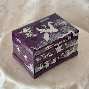 May include: A rectangular, deep purple jewelry box with two drawers and a hinged lid. The box features white inlaid mother-of-pearl designs of cranes and trees. The drawer pulls are silver-coloured and butterfly-shaped. The box is approximately 20 cm long.