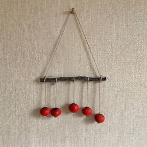 May include: A rustic wall hanging featuring a dark wooden branch suspended by twine. Six bright red, round ornaments hang from the branch by twine. The wall behind the hanging is a textured, light beige color.