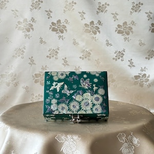 May include: A green jewelry box with a floral and butterfly design. The box has a silver latch.