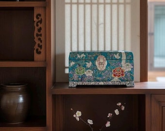 May include: A rectangular storage box with a teal background featuring intricate floral and bird patterns in shades of red, white, and green. The box has a metallic latch on the front and sits on a wooden shelf next to a small green vase with flowers.