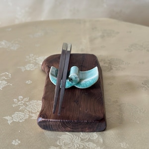 May include: A wooden chopstick rest with a teal ceramic bird-shaped holder and dark brown chopsticks. The rest is a dark brown, rectangular piece of wood with a natural edge. The chopsticks are resting on the bird-shaped holder.