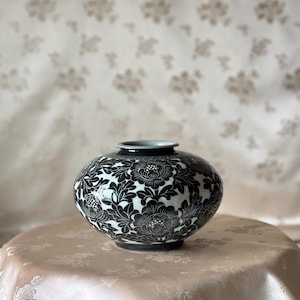 May include: A round ceramic vase featuring a high-contrast black and white floral pattern. The surface is decorated with detailed, repeating flower and leaf motifs. The vase has a short, narrow neck with a smooth, light-colored rim opening.