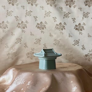 May include: A pale mint green ceramic container shaped like a traditional East Asian building with a tiered, tiled roof. The object features a smooth, cylindrical base and a removable lid with detailed ridges mimicking roof tiles and upturned corners.