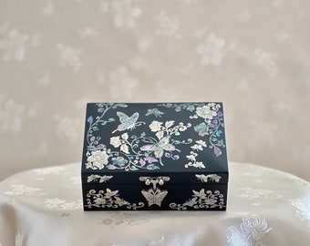 Pode incluir: Uma caixa de madeira preta com tampa articulada decorada com um design floral e de borboletas de madrepérola. A caixa tem um fecho de metal prateado.