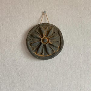 May include: A round, dark gray decorative wall hanging with a floral design. The center of the flower is a lighter shade of brown. The wall hanging is suspended by a thin, natural-colored cord.