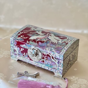 May include: A burgundy jewelry box with a crane design inlaid with iridescent mother-of-pearl. The box has a silver clasp and feet, and a matching tassel. The box is approximately 6 inches long, 4 inches wide, and 3 inches tall.