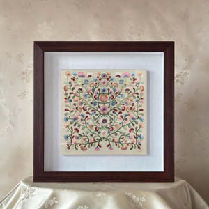 Può includere: Ricamo floreale incorniciato con una cornice in legno marrone scuro. Il ricamo presenta un disegno simmetrico di fiori e viti colorati nelle tonalità del rosa, blu, verde e rosso su sfondo crema. La cornice è quadrata.