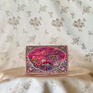 May include: A pink jewelry box with a floral design. The box has a rectangular shape with a curved top and is decorated with intricate floral and butterfly patterns in various colours. The box is sitting on a white surface.
