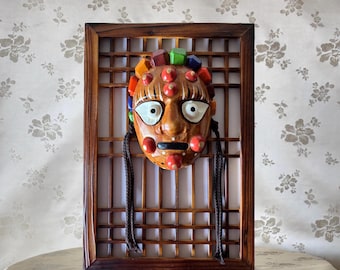 May include: A traditional Korean mask with a colorful face and a wooden frame. The mask is decorated with red, orange, yellow, green, blue, and purple beads. The mask is hanging on a wooden lattice.