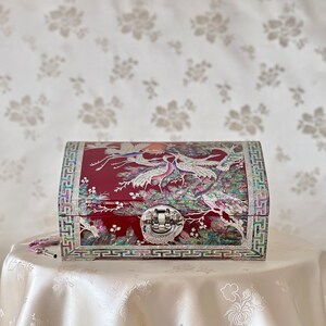 May include: A red wooden jewellery box with a hinged lid and a silver latch. The box is decorated with a mother-of-pearl inlay of cranes and pine trees.