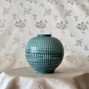 May include: A teal ceramic vase with a round body and a narrow neck. The vase features vertical ribbed detailing and a decorative pattern around the middle. It sits on a light-coloured surface against a floral patterned backdrop.