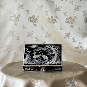 May include: A black wooden box with a hinged lid decorated with a mother-of-pearl inlay of cranes and a pine tree. The box has a silver latch.