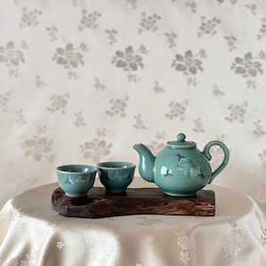 May include: A teal ceramic tea set with a teapot and two cups, resting on a dark wooden tray. The teapot has a curved spout and handle, with a lid. The cups are small and round. The background is a floral patterned wallpaper.