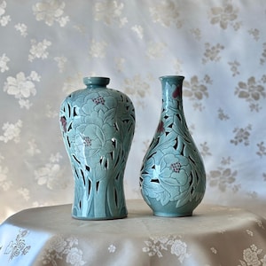 Puede incluir: Dos jarrones de cerámica con intrincados diseños florales. Los jarrones son de color azul claro con patrones florales blancos y rojos. Los jarrones están sobre una superficie azul claro.