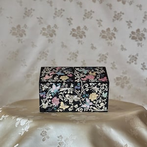 Puede incluir: Una caja decorativa negra con tapa con bisagras, adornada con mariposas coloridas y diseños florales. La caja presenta detalles intrincados y un acabado brillante, descansando sobre una superficie color crema.