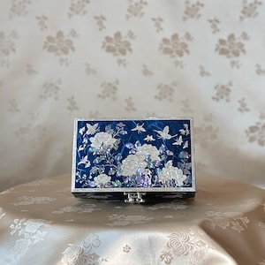 May include: A rectangular, dark blue jewelry box with a floral and butterfly design. The box is inlaid with mother-of-pearl, featuring white flowers and butterflies. The box has a silver clasp and is set on a patterned surface.